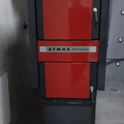 Czerwono-czarny piec marki Atmos Drevoplyn w kotłowni, widoczne rury i zbiornik wyr&oacute;wnawczy.