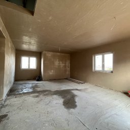 Tom Tynk - Wnętrze w trakcie prac wykończeniowych, ściany i sufit pokryte świeżym tynkiem w odcieniach beżu, widoczne ślady wilgoci na podłodze, puszki z farbą w rogu pomieszczenia i fragment instalacji...