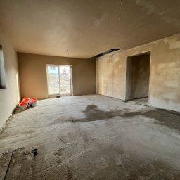 Tom Tynk - Pokój w trakcie prac wykończeniowych: ściany pokryte świeżym tynkiem, na podłodze widoczne ślady zaprawy i kilka czerwonych wiader z materiałem. Widoczne niedoskonałości na ścianach i podłodze.
