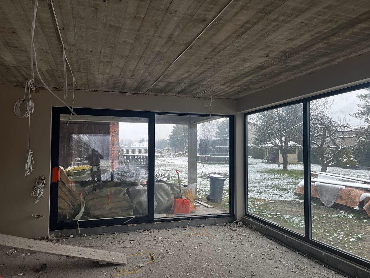 Surowy betonowy sufit w nowym domu z dużymi oknami, widoczne kable zwisające ze stropu, worki z materiałem izolacyjnym i czerwona łopata w tle za oknem, na zewnątrz widok na ośnieżony ogród.