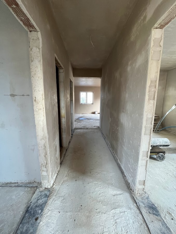 Korytarz w trakcie prac wykończeniowych, świeżo nałożone tynki na ścianach, widoczne surowe wykończenie ościeżnic i podłogi, w tle okno z widokiem na zieleń.