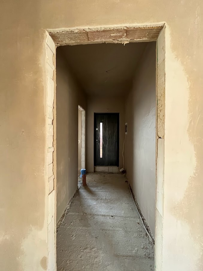 Korytarz w trakcie prac wykończeniowych; ściany pokryte świeżym, jasnym tynkiem, widoczne fragmenty surowej cegły nad ościeżnicą, rury instalacyjne na podłodze.