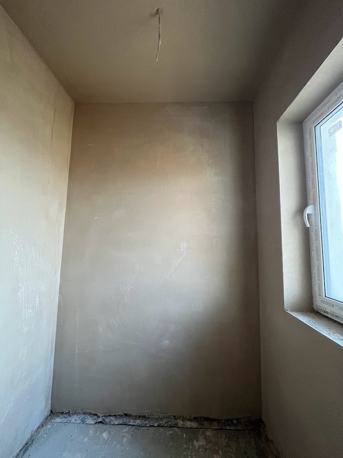 Świeżo położona, jasna, gładka warstwa tynku na ścianach i suficie w pomieszczeniu z oknem. Widoczne niedoskonałości przy podłodze i wystający kabel z sufitu.