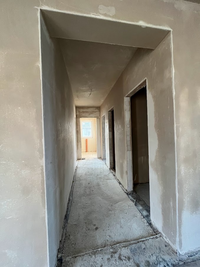 Korytarz w trakcie prac wykończeniowych, świeżo położone tynki na ścianach i suficie, widoczne futryny drzwiowe bez drzwi, podłoga przygotowana do dalszych prac.