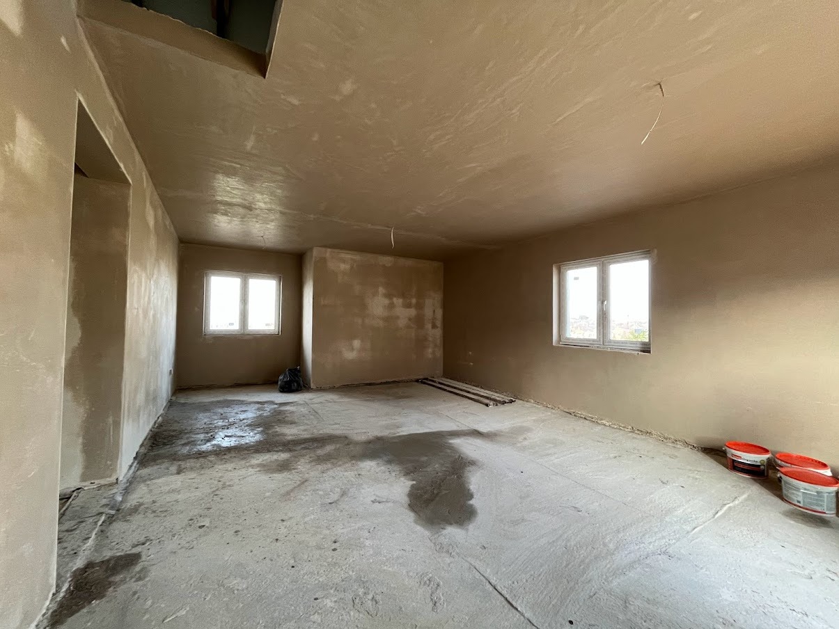 Wnętrze w trakcie prac wykończeniowych, ściany i sufit pokryte świeżym tynkiem w odcieniach beżu, widoczne ślady wilgoci na podłodze, puszki z farbą w rogu pomieszczenia i fragment instalacji...