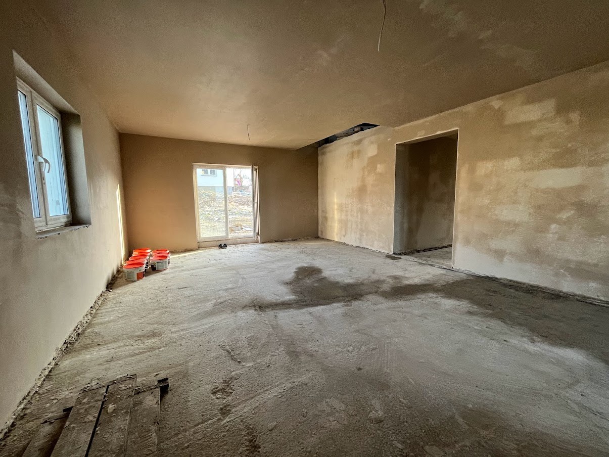 Pokój w trakcie prac wykończeniowych: ściany pokryte świeżym tynkiem, na podłodze widoczne ślady zaprawy i kilka czerwonych wiader z materiałem. Widoczne niedoskonałości na ścianach i podłodze.