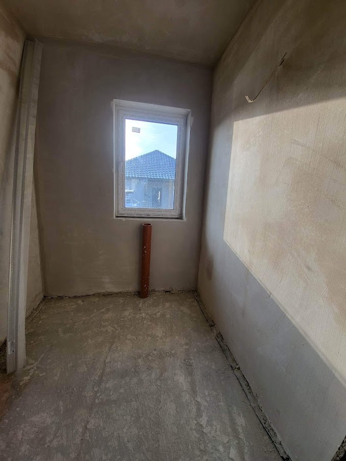 Surowe wnętrze pomieszczenia po tynkowaniu maszynowym ścian, widoczne okno z widokiem na dach budynku, pionowa rura i metalowy profil przy ścianie.