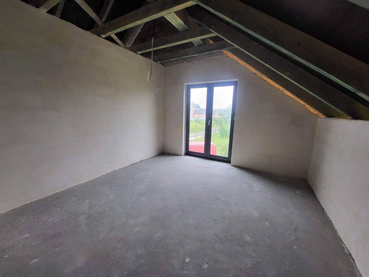 Pokój na poddaszu po nałożeniu tynków, widoczne drewniane elementy konstrukcji dachu i okno balkonowe z widokiem na zewnątrz.