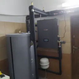 Szary zbiornik buforowy i jednostka grzewcza z czarnymi rurami izolacyjnymi w pomieszczeniu z beżowymi płytkami. Widoczny biały zbiornik wyr&oacute;wnawczy i logo 'HEATECO' na jednostce grzewczej.