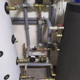 Instalacja grzewcza z białym i grafitowym zasobnikiem, rurami w szarej izolacji, zaworami i pompą obiegową, widoczne połączenia miedziane i złączki.