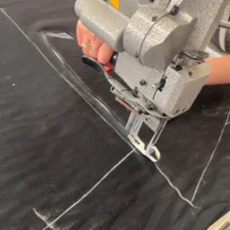 Elektryczny nóż krawiecki w dłoni tnie czarny materiał po białych liniach. Widoczny fragment stołu krawieckiego i maszyny.