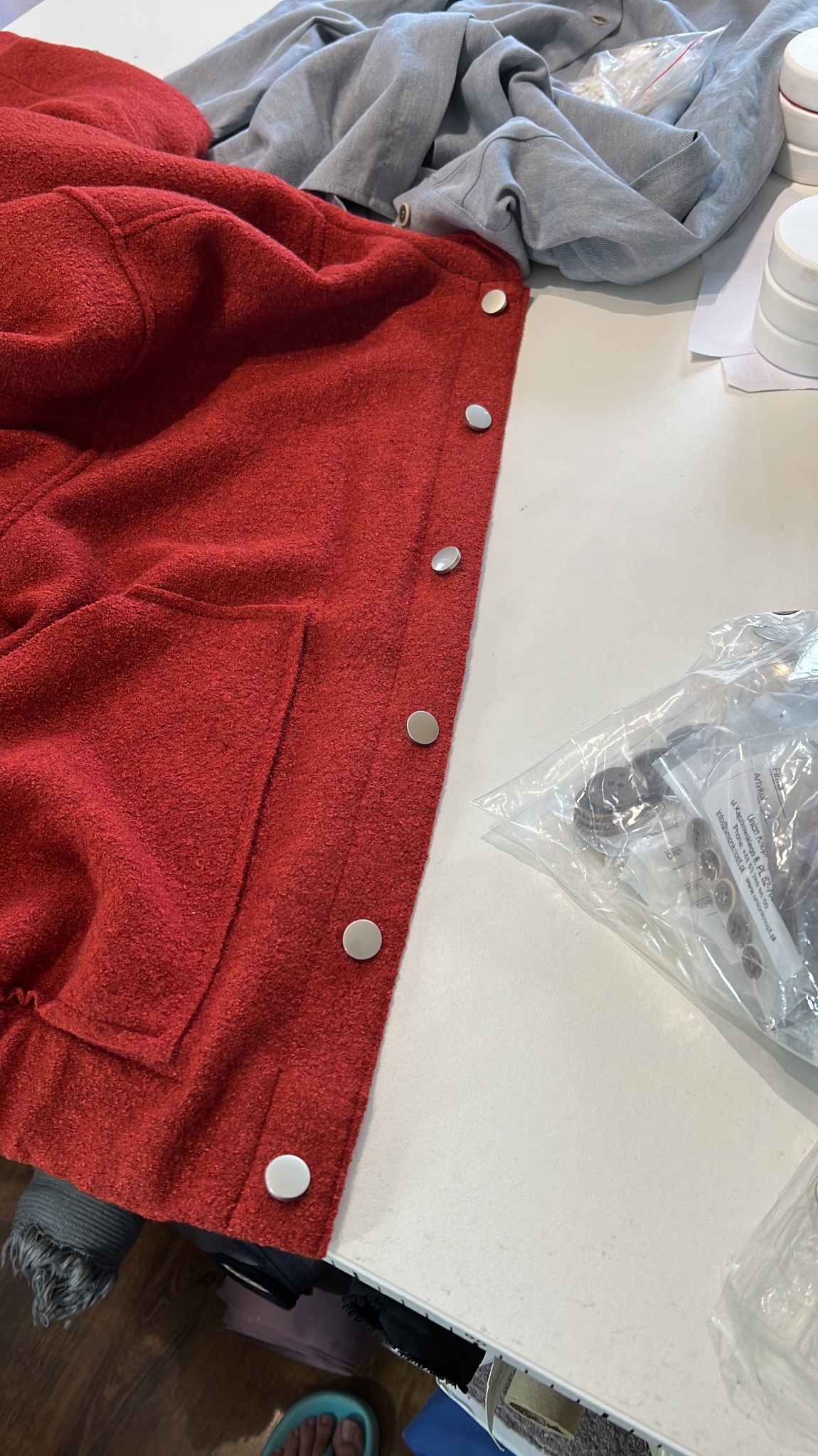 Czerwona kurtka z kieszenią i srebrnymi guzikami leżąca na stole obok worka z zapasowymi guzikami i szarych ubrań.