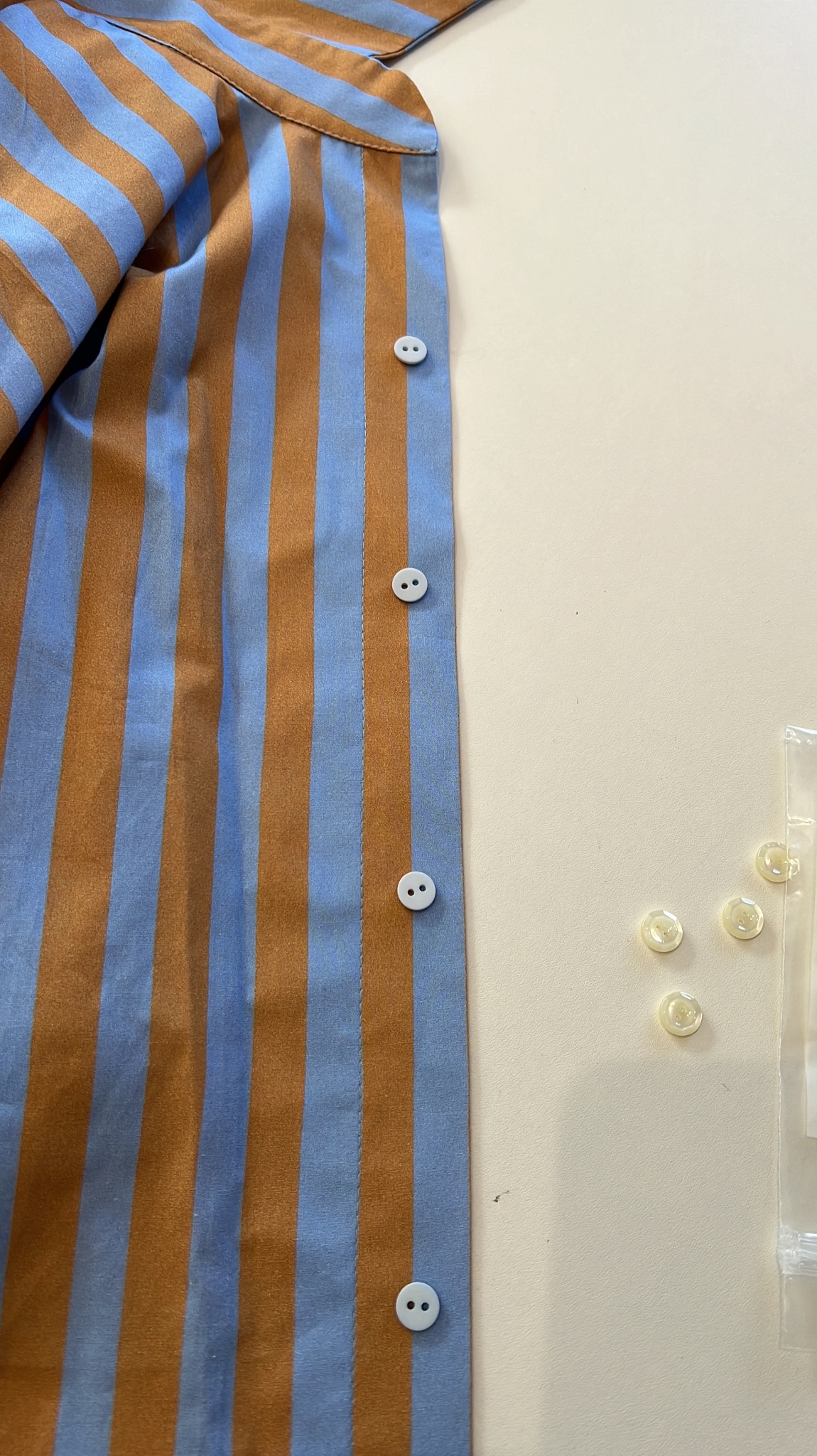Fragment koszuli w pionowe, brązowo-niebieskie pasy z przyszytymi białymi guzikami, obok leżą dodatkowe guziki w plastikowej torebce.