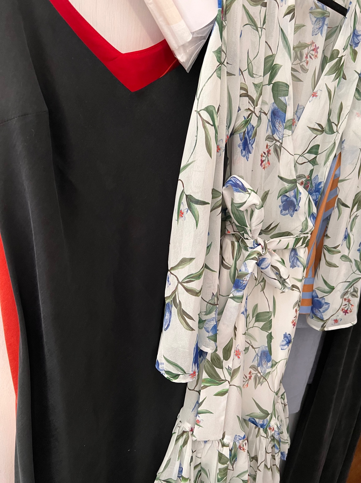 Ujęcie z bliska wiszących ubrań: czarna sukienka z czerwonym dekoltem i zwiewna bluzka w kwiaty z wiązaniem w pasie.