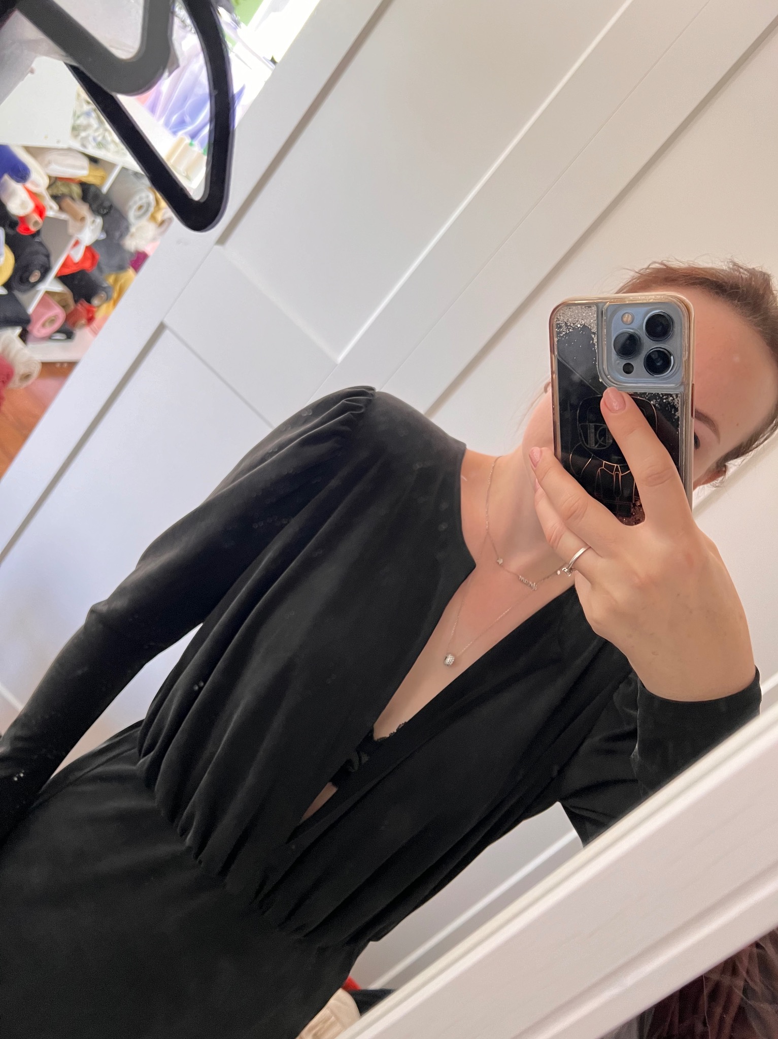 Kobieta robi selfie w lustrze, ubrana w czarną sukienkę z długim rękawem i dekoltem w serek, widoczny fragment garderoby z tkaninami w tle.