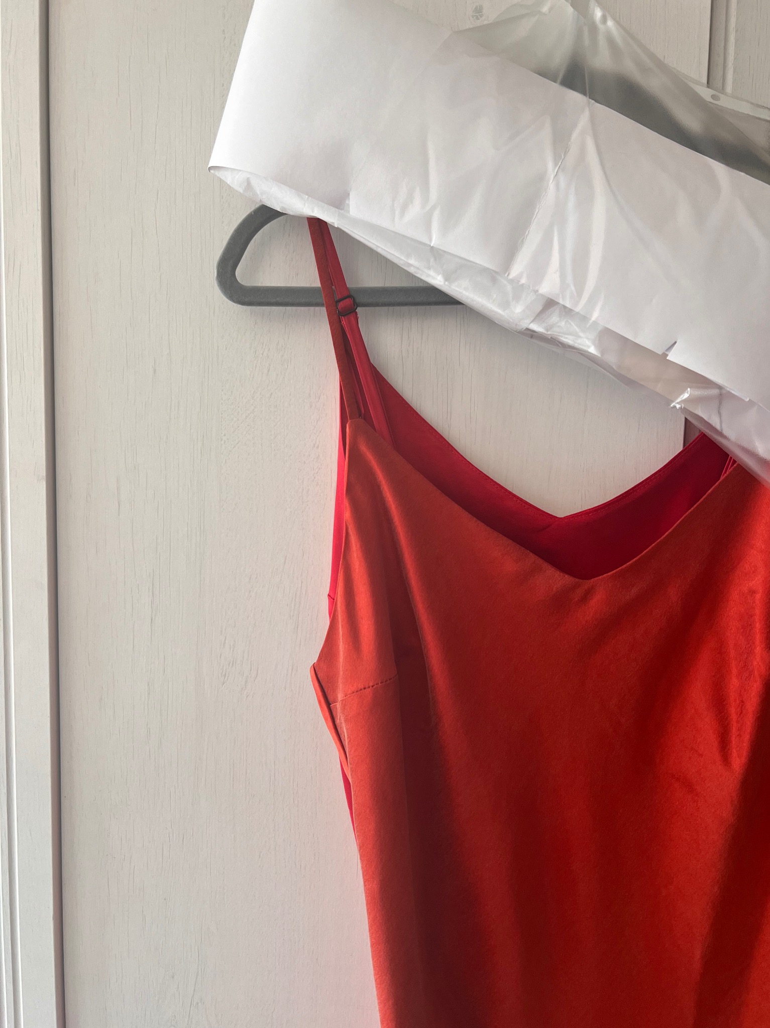 Czerwona sukienka na ramiączkach, wisząca na szarym wieszaku z białym pokrowcem ochronnym na tle białych drzwi.