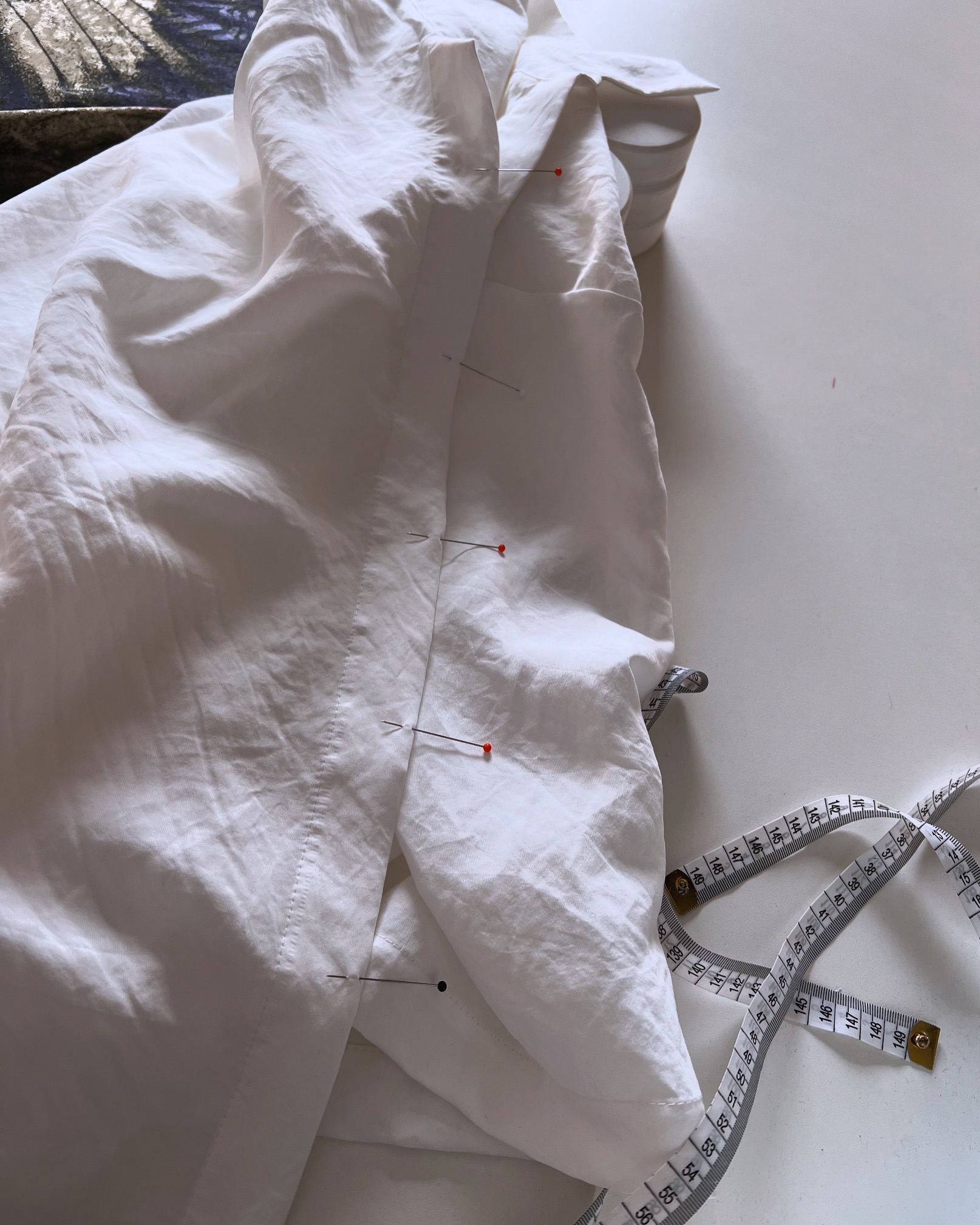 Biała, lniana koszula z widocznymi szpilkami i miarką krawiecką na białym tle, sugerująca etap dopasowywania odzieży.