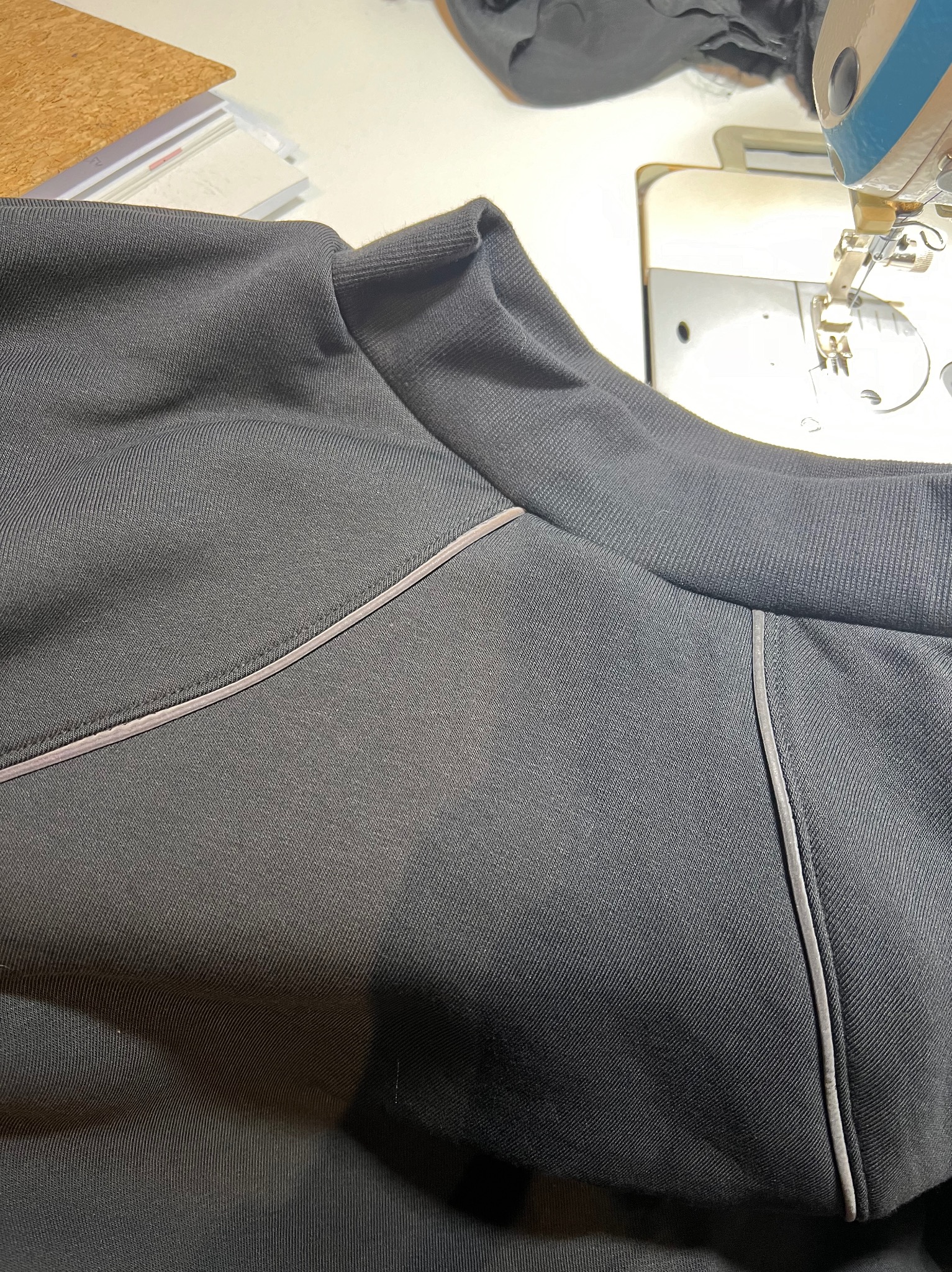 Szara bluza z kontrastową lamówką na dekolcie, widoczna maszyna do szycia w tle, ukazująca etap produkcji odzieży.