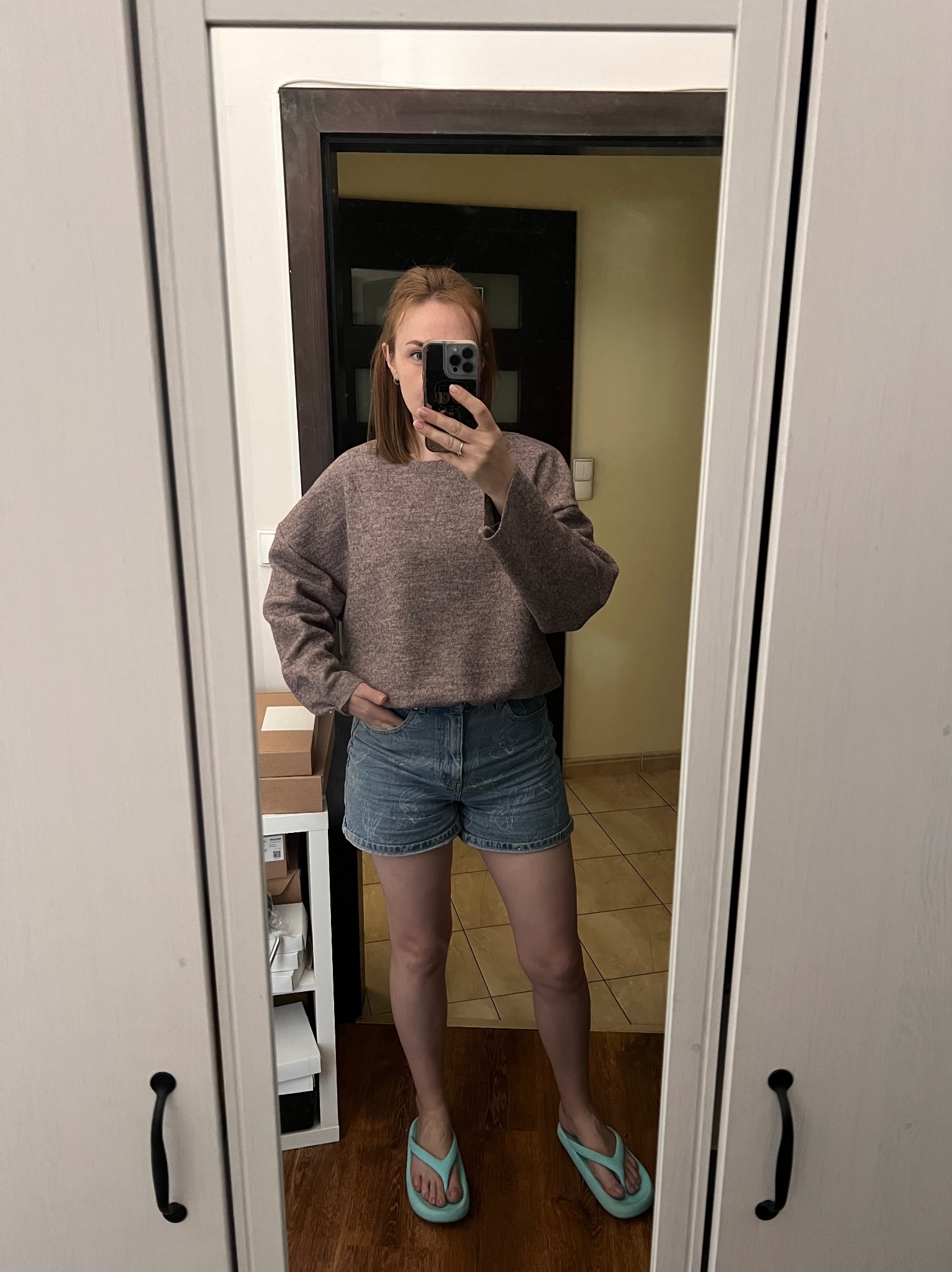 Osoba robiąca selfie w lustrze, ubrana w szary sweter o grubym splocie i dżinsowe szorty, z widoczną półką z pudełkami w tle.