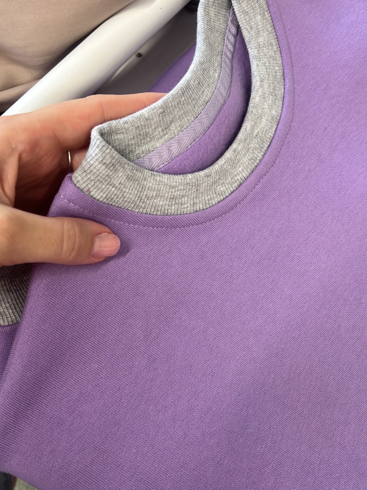 Liliowa bluza dresowa z szarym ściągaczem, trzymana w dłoni, ukazująca detale wykończenia dekoltu i jakość materiału. Widoczny fragment ubrania na wieszaku w tle.
