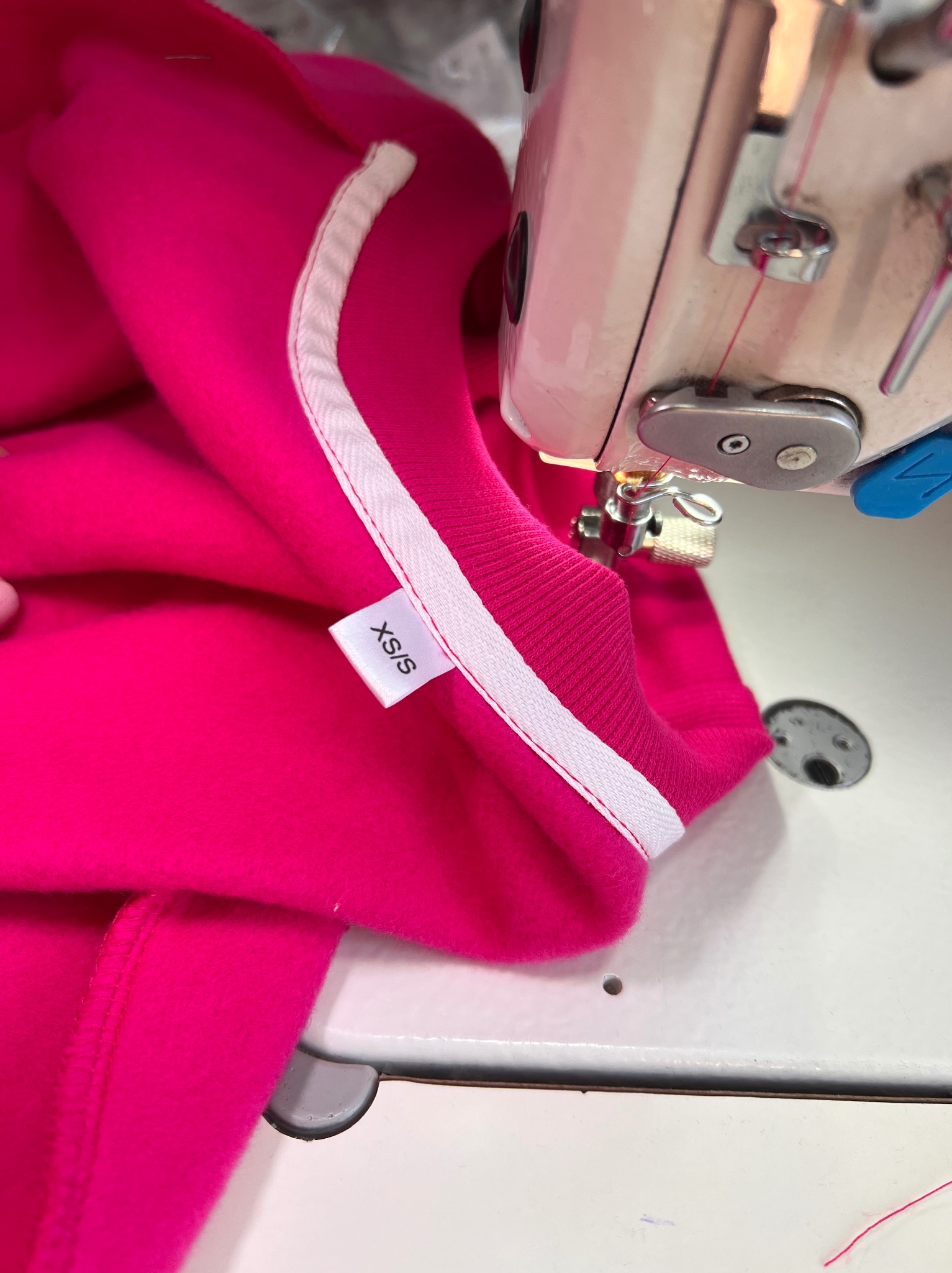 Różowa bluza z metką XS/S pod igłą maszyny do szycia, zbliżenie na etap produkcji odzieży w szwalni. Widoczna biała lamówka.