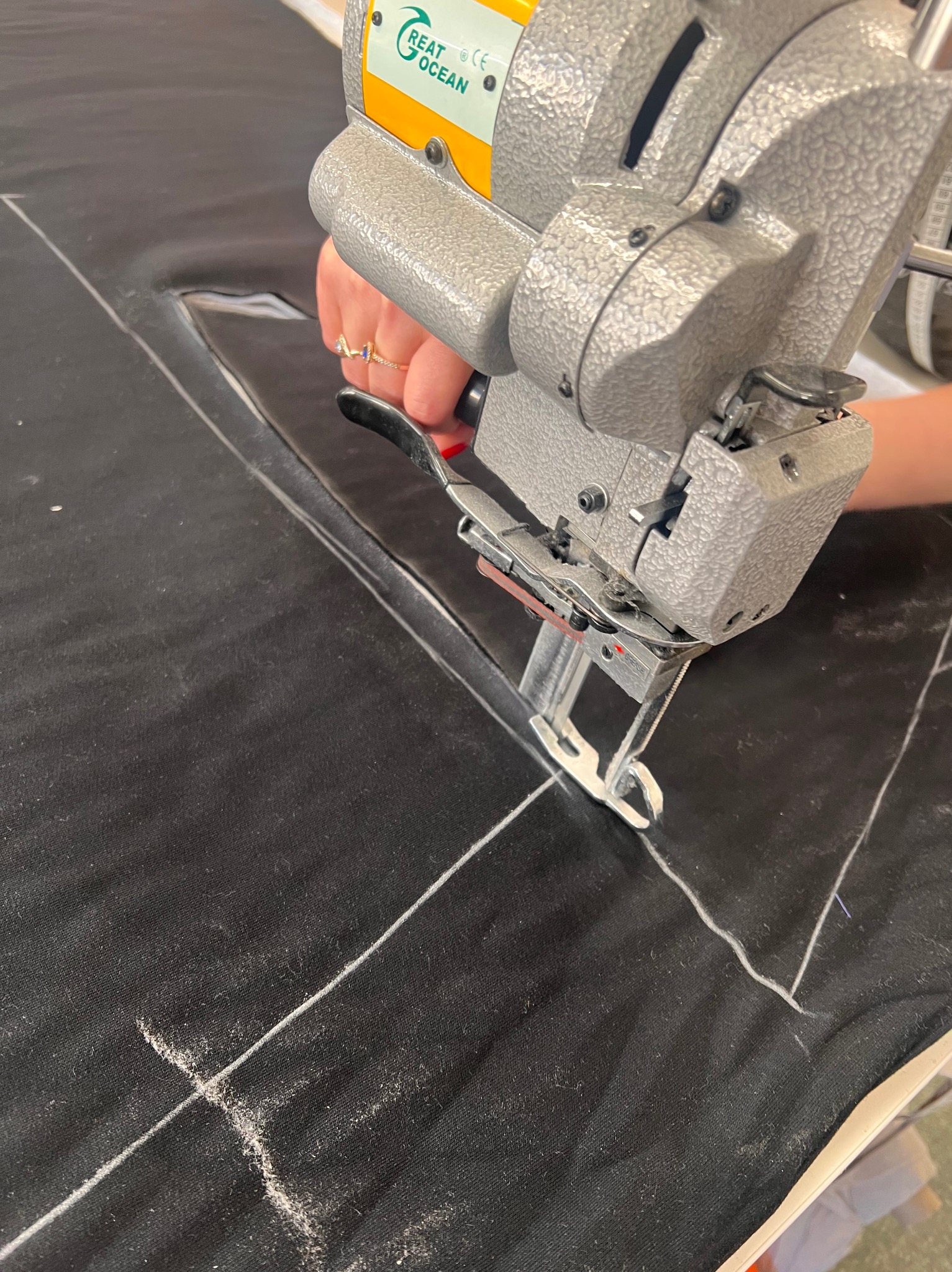 Elektryczny nóż krawiecki w dłoni tnie czarny materiał po białych liniach. Widoczny fragment stołu krawieckiego i maszyny.