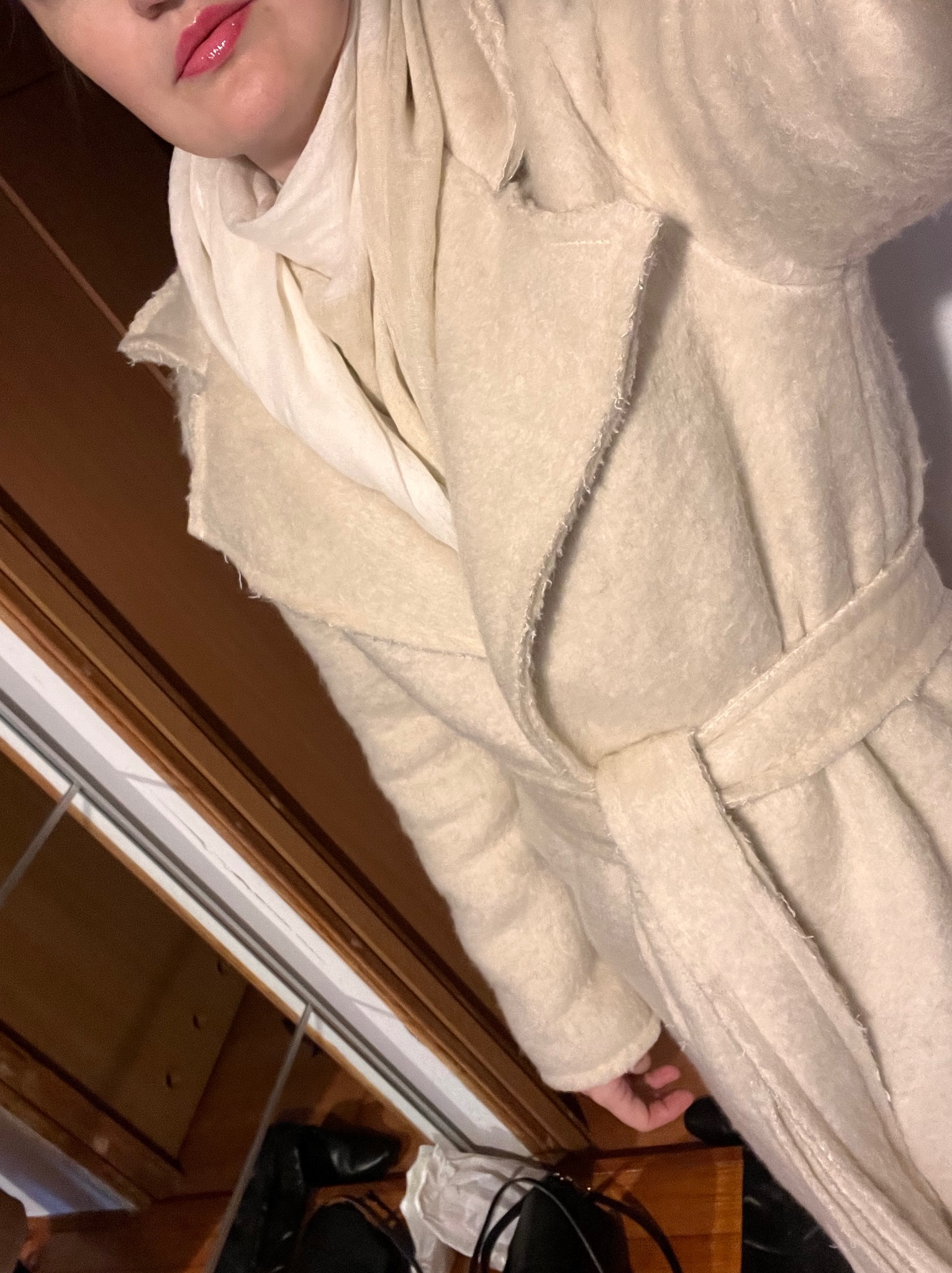 Kremowy, wiązany płaszcz z szerokim kołnierzem, założony na białą bluzkę i szal. Widoczny fragment twarzy i dłoni. Ujęcie z góry, lekko obrócone.