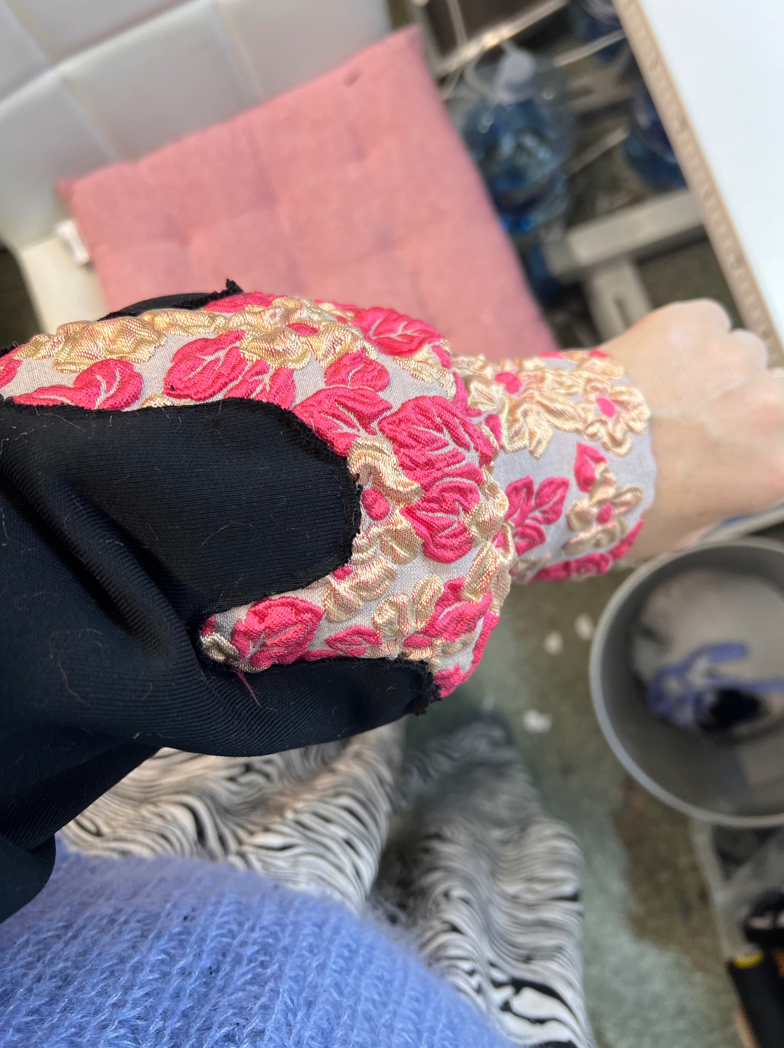 Fragment rękawa czarnej sukienki z ozdobnym haftem w kwiaty w odcieniach różu i złota, widoczny na tle wzorzystych spodni i niebieskiego swetra.