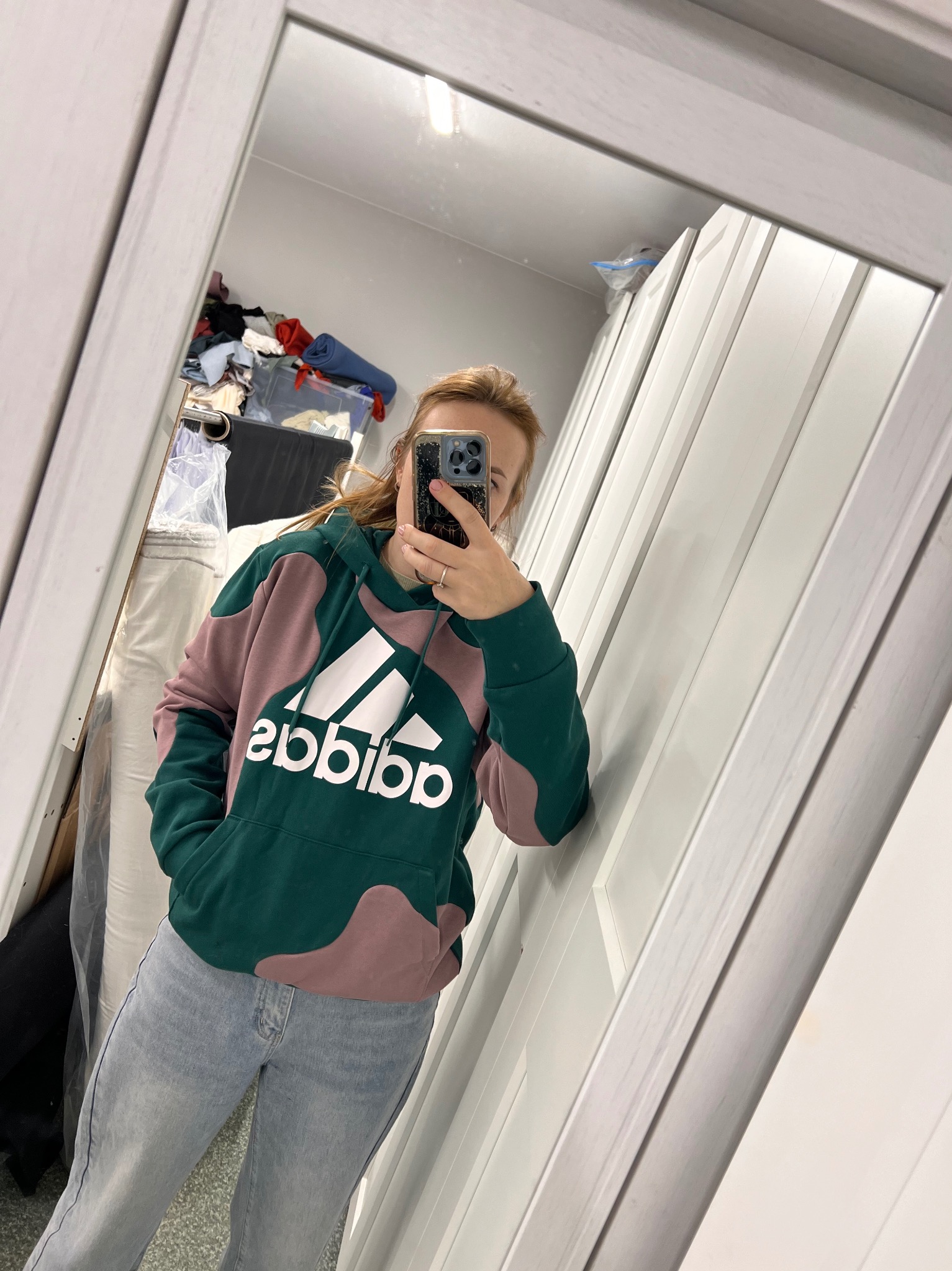 Osoba w lustrze prezentuje zielono-różową bluzę Adidas. W tle widoczne materiały, sugerujące szwalnię. Uwagę zwraca nietypowe połączenie kolorów bluzy.