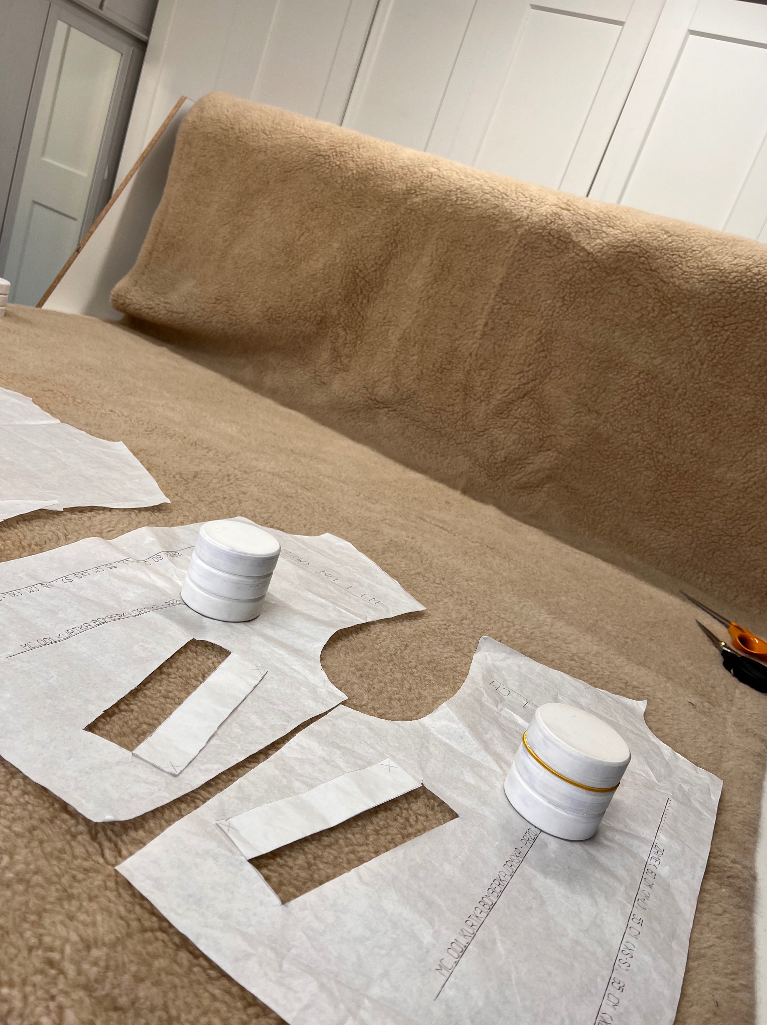 Rozłożony na beżowym futrze szablon odzieży z obciążnikami, przygotowany do krojenia. W tle białe szafy.