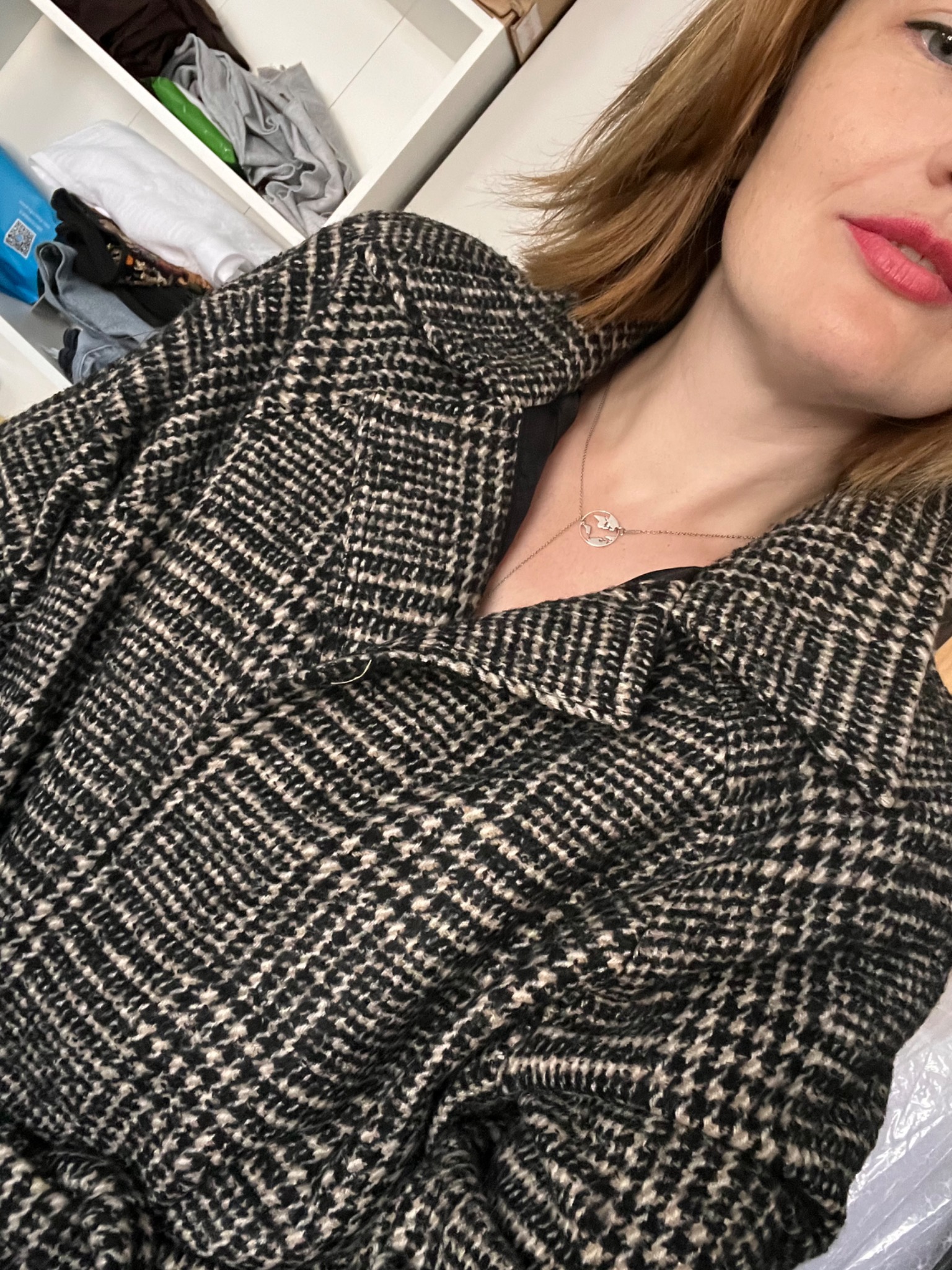 Kobieta w kurtce w pepitkę z widocznym dekoltem i srebrnym naszyjnikiem. W tle białe półki z ubraniami. Ujęcie z bliska, skupione na detalu tkaniny i biżuterii.