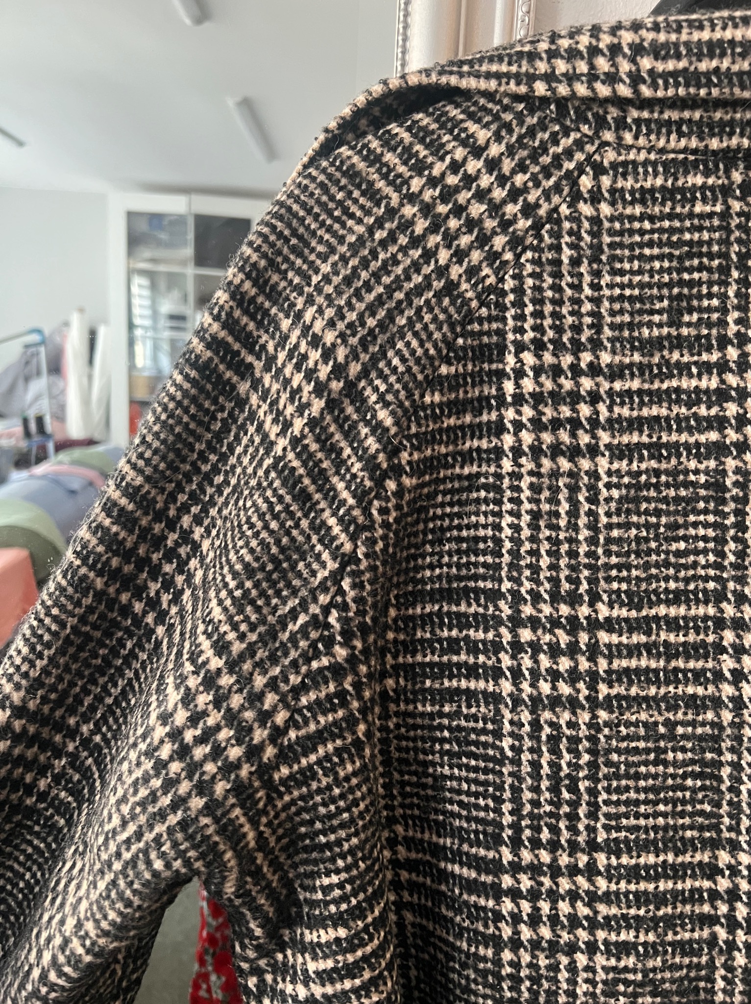 Fragment wełnianego płaszcza w pepitkę, widoczny rękaw i kołnierz. W tle jasne pomieszczenie z wyposażeniem szwalni, podkreślające rzemieślniczy charakter.