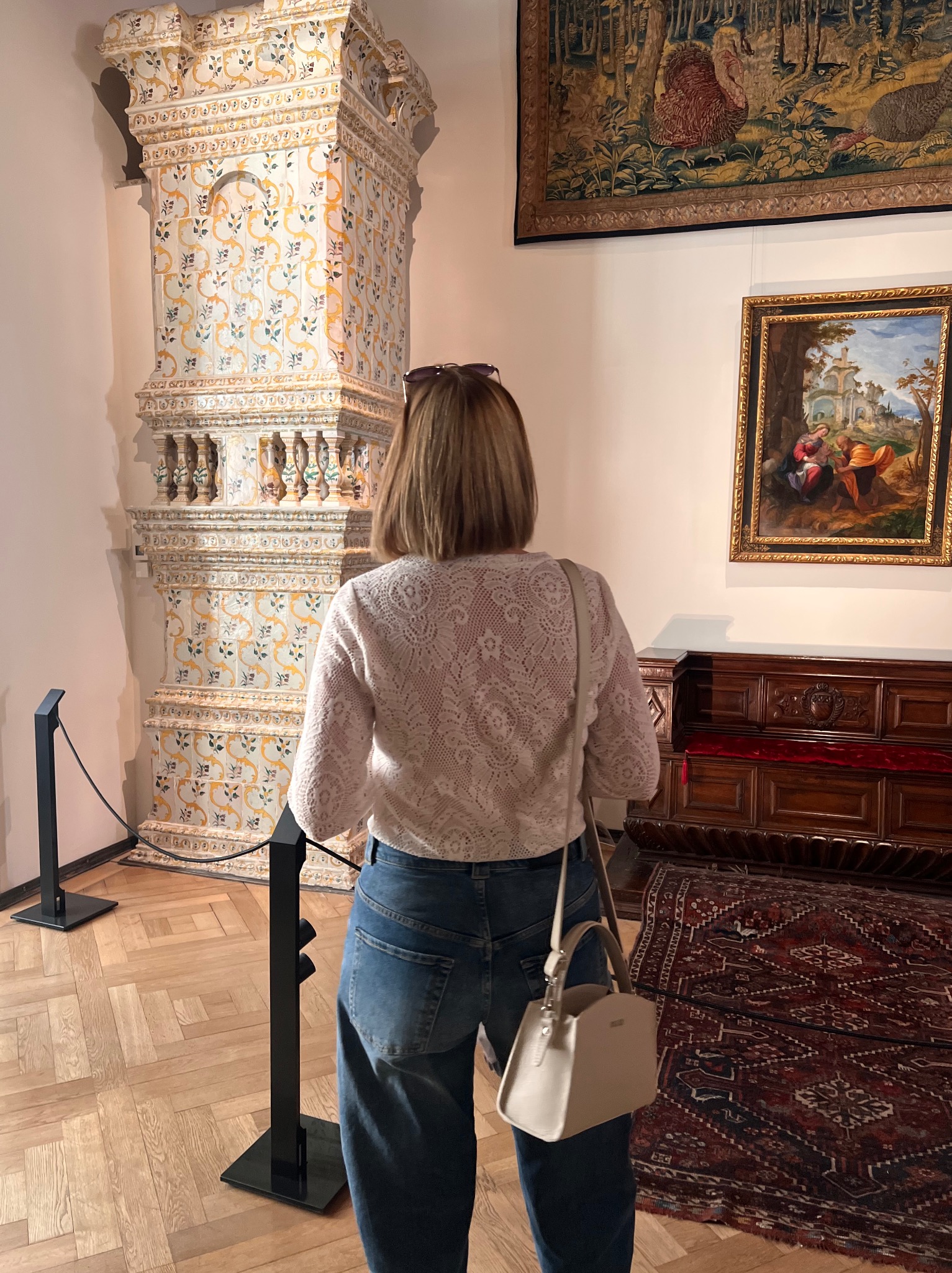 Kobieta w koronkowej bluzce ogląda zdobiony, kaflowy piec w muzeum. W tle obrazy i dywan. Uwagę przykuwa bogate zdobienie pieca i wzór bluzki.