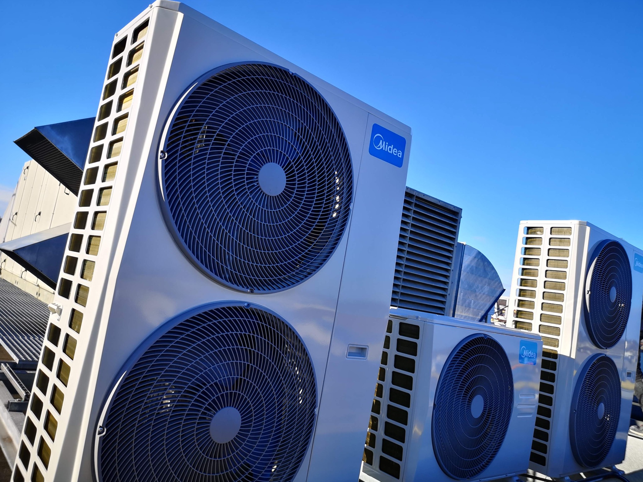 Instalacja kilku jednostek zewnętrznych klimatyzacji marki Midea na dachu budynku, widok z dołu na tle błękitnego nieba.