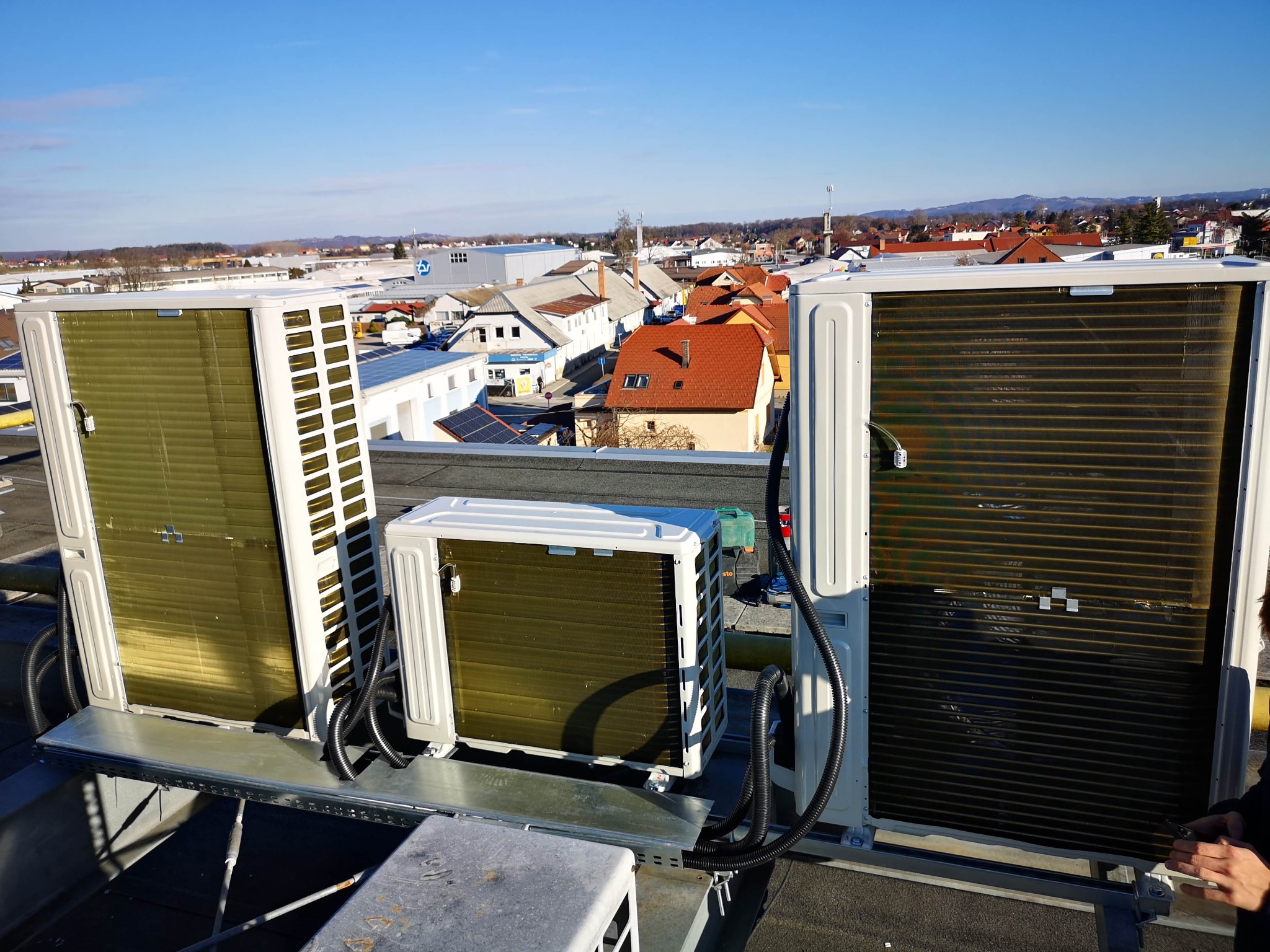 Trzy jednostki zewnętrzne klimatyzacji na dachu budynku z widokiem na miasto w tle, pod błękitnym niebem.