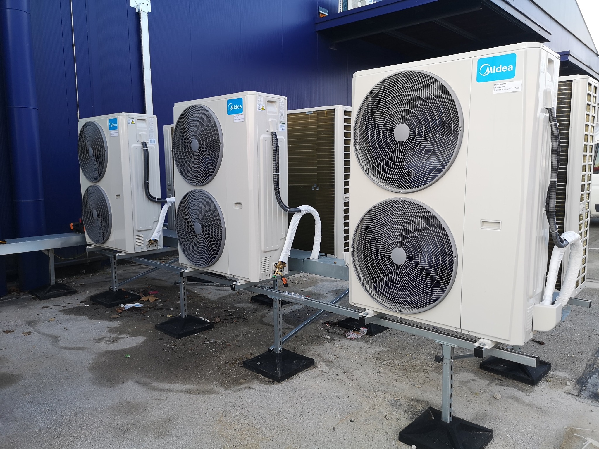 Rząd zewnętrznych jednostek klimatyzacyjnych Midea, ustawionych na metalowych konstrukcjach na zewnątrz budynku z niebieską elewacją. Widoczne przewody i elementy montażowe.