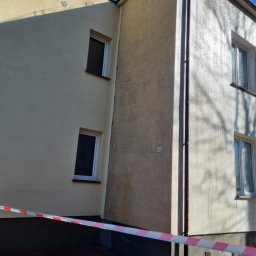 Pro Clean Cezary Woźniak - Dwupiętrowy budynek z elewacją w odcieniach beżu, częściowo umytą, z widocznymi zaciekami. Czerwono-biała taśma zabezpieczająca u dołu. Błękitne niebo w tle.