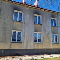 Pro Clean Cezary Woźniak - Dwupiętrowy budynek z elewacją z widocznymi zabrudzeniami i zaciekami. Przy ścianie oparta drabina. Widoczne okna z białymi ramami i fragment niebieskiego nieba.