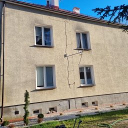 Pro Clean Cezary Woźniak - Dwupiętrowy budynek z beżową elewacją w słoneczny dzień. Widoczne okna z białymi ramami, rynna i fragment ogrodu z zielonym trawnikiem i roślinami.