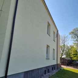Pro Clean Cezary Woźniak - Jasna elewacja budynku z ciemną rynną, widok z dołu w słoneczny dzień. Widoczne okna i fragment trawnika. Budynek po renowacji, z nową, jednolitą fakturą.