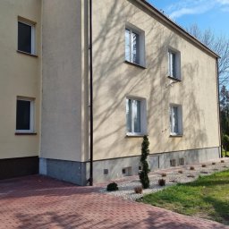 Pro Clean Cezary Woźniak - Dwupiętrowy budynek z beżową elewacją, widoczny z perspektywy chodnika z czerwonej kostki. Na elewacji widoczne cienie drzew i małe okna piwniczne.