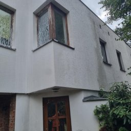 Pro Clean Cezary Woźniak - Biała elewacja domu po myciu, z zaciekami na oknach i ścianach. Widoczne okna z drewnianą ramą i fragment zieleni. Część ściany z cegły. Pochmurne niebo w tle.