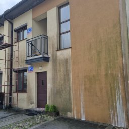 Pro Clean Cezary Woźniak - Budynek z widocznymi zabrudzeniami na elewacji, częściowo umyty fragment ściany. Na pierwszym planie kostka brukowa i fragment rusztowania.