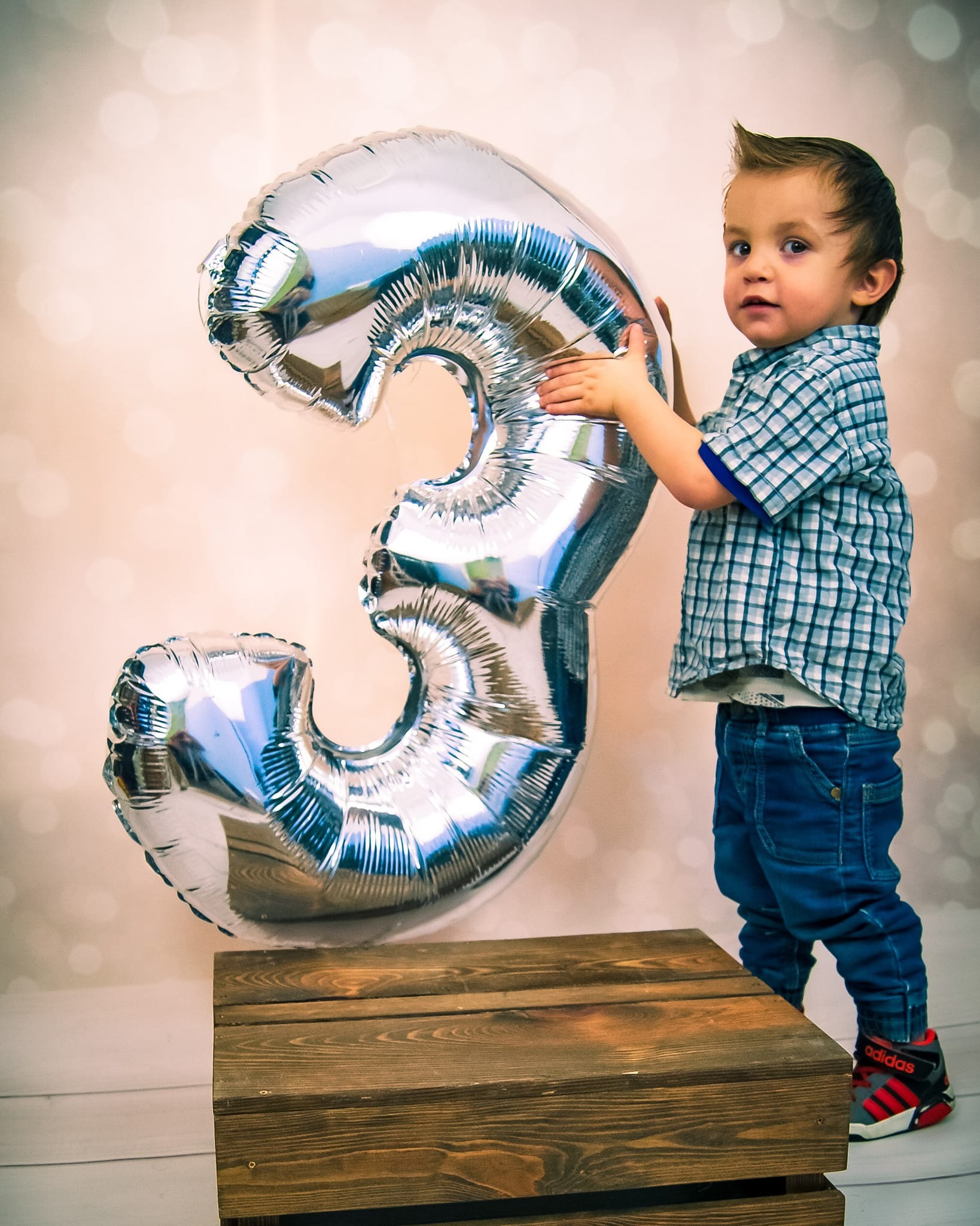 Trzyletni chłopiec w kraciastej koszuli i jeansach trzyma duży, srebrny balon w kształcie cyfry 3, stojąc obok drewnianej skrzyni na tle jasnego, rozmytego tła.