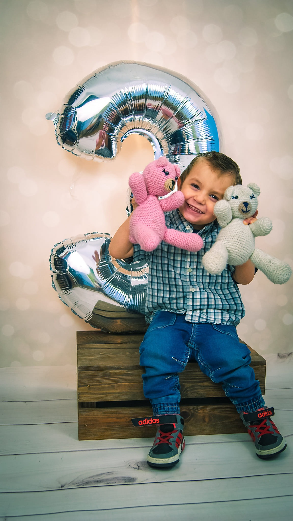 Uśmiechnięty chłopiec siedzi na drewnianej skrzyni, trzymając w rękach dwa pluszowe misie, różowego i białego, z balonem w kształcie cyfry trzy w tle.