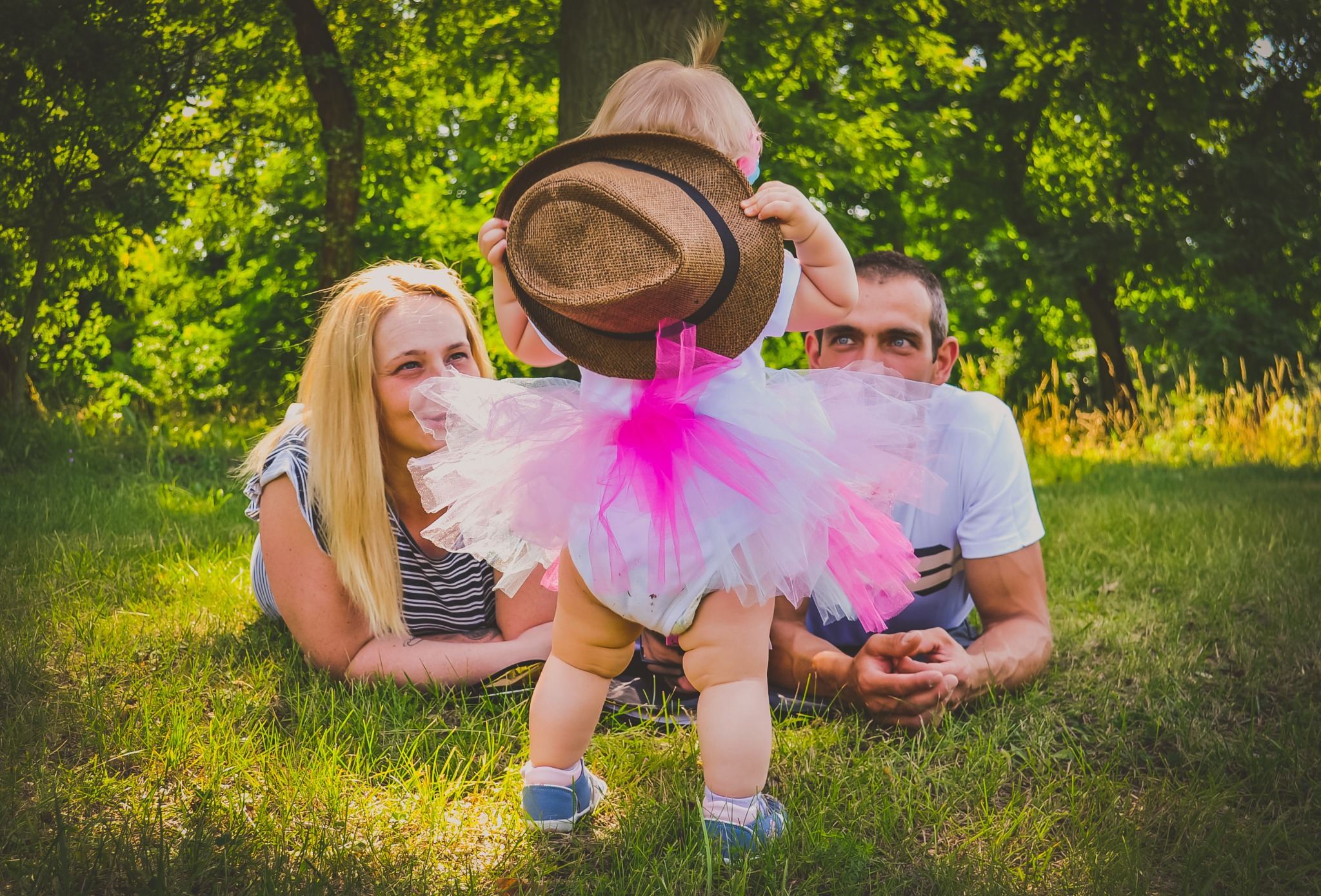 Rodzina leżąca na trawie: blondwłosa mama i tata z niebieskimi oczami obserwują małą córeczkę w różowej tutu, która trzyma na twarzy brązowy kapelusz.