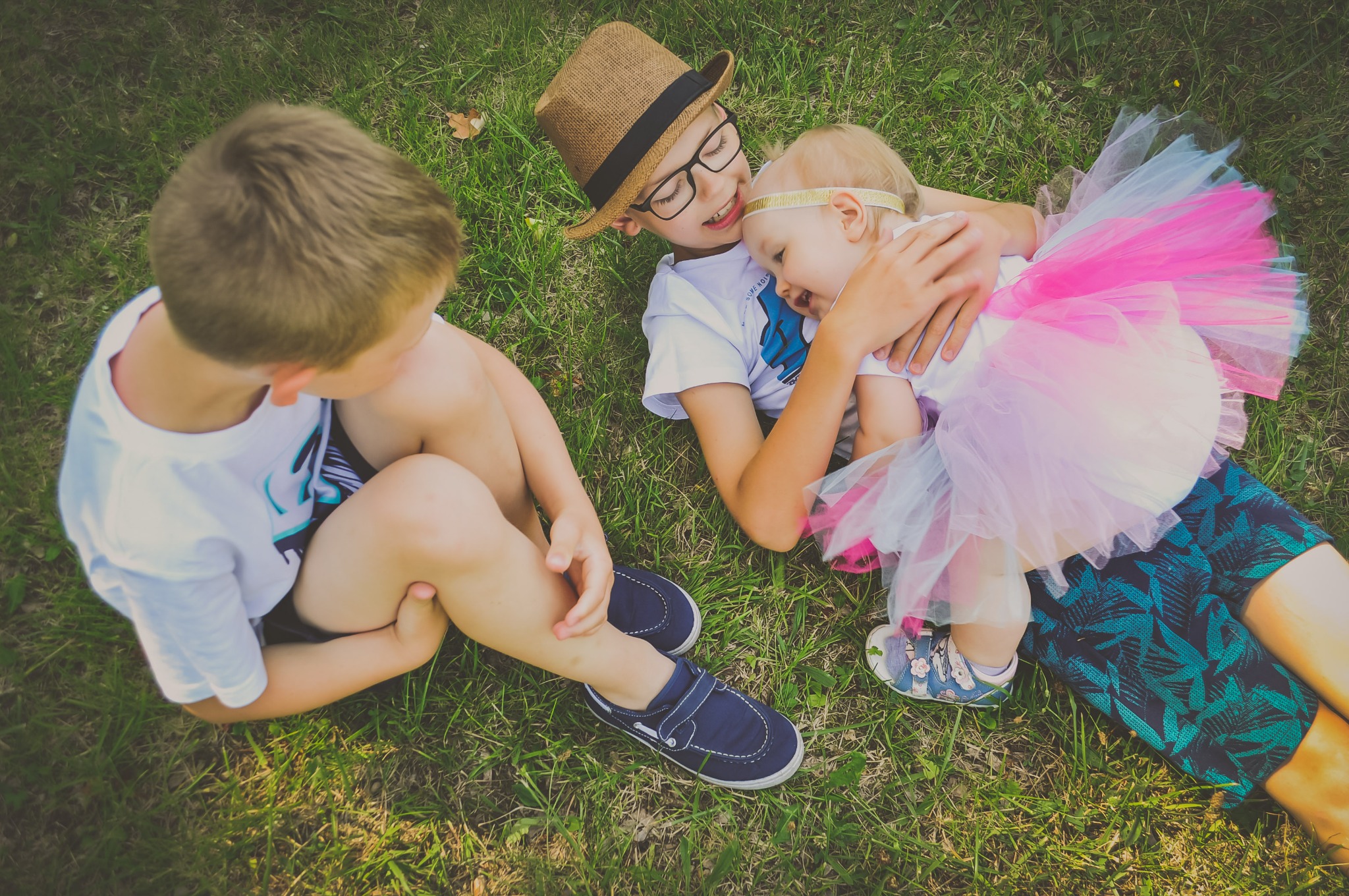 Dwóch chłopców i dziewczynka leżą na trawie: jeden chłopiec w kapeluszu i okularach obejmuje dziewczynkę w tiulowej spódniczce, drugi chłopiec siedzi obok.