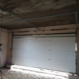 Wnętrze garażu w trakcie montażu segmentowej bramy garażowej w kolorze białym, widoczny napęd sufitowy, surowy betonowy strop i ściany z pustaków.