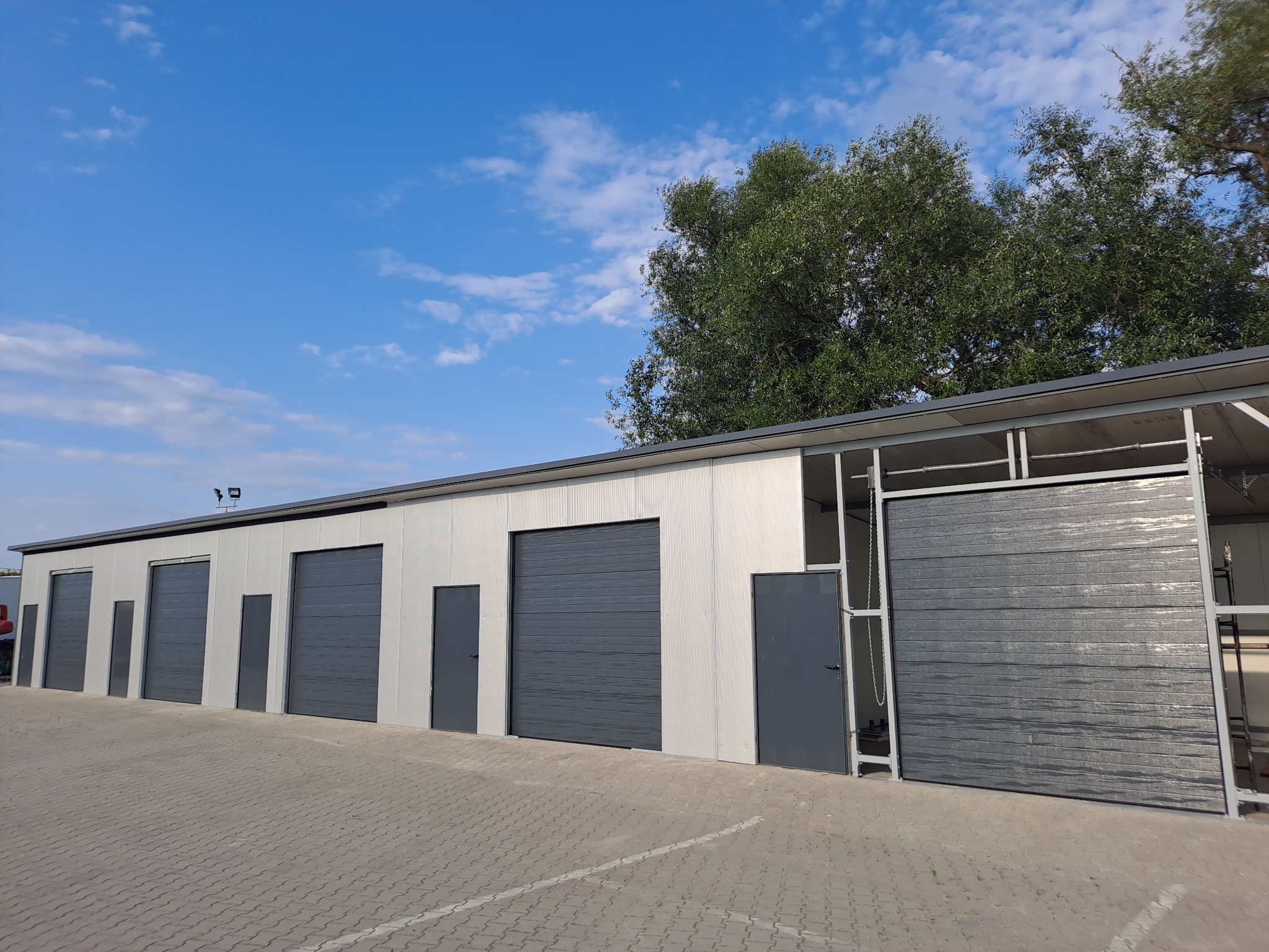 Rząd szarych bram garażowych segmentowych z drzwiami wejściowymi w budynku magazynowym z blachy trapezowej na tle błękitnego nieba z drzewami.