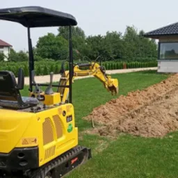 Żółta mini koparka na trawniku w trakcie wykonywania wykopu pod fundament domu w stanie surowym otwartym.